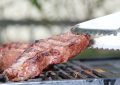 Beef steak cooking on a barbecue grill with a serrated metal tongs lifting the edge