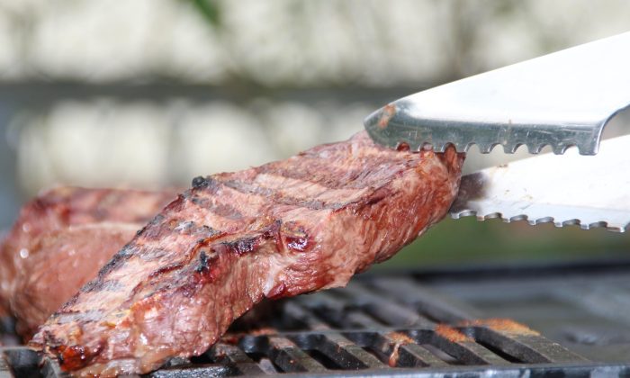 Beef steak cooking on a barbecue grill with a serrated metal tongs lifting the edge