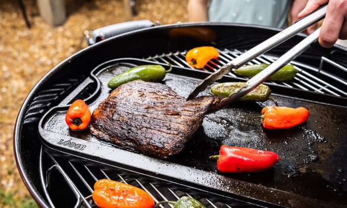Beef steak grilling on a barbecue with colorful peppers nearby, tongs lifting the meat outdoors.