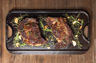 Two seared beef steaks with garlic and rosemary on a Lodge cast-iron skillet over a wooden table.