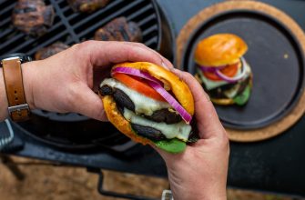 Person grips a cheeseburger with melted cheese, tomato, red onion, and lettuce over a grill.