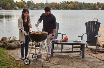 Couple grilling outdoors by a lake with fall foliage in the background, a round charcoal grill in use.