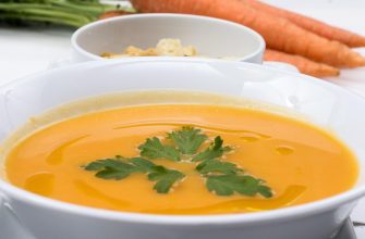 Creamy orange carrot soup in a white bowl with parsley garnish, croutons in the background and fresh carrots nearby.