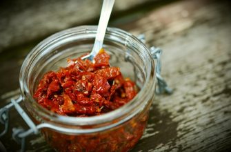 Open glass jar filled with chopped red peppers; a spoon rests inside on a weathered wooden surface.