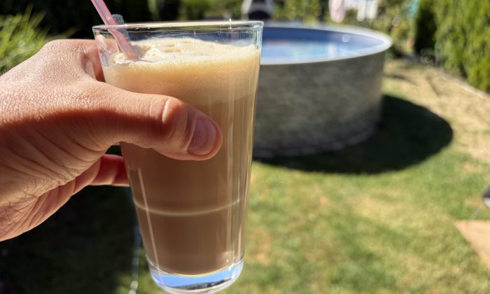 Hand holding a tall glass of iced coffee with a pink straw, outdoors near a pool under sunny skies.