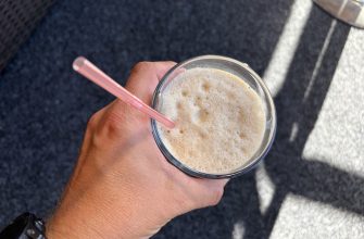 Hand holding a glass of frothy coffee with a pink straw, over a dark textured surface outside.